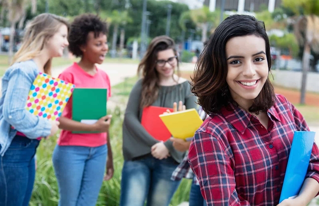 A student in a plaid shirt smiling at the camera, with classmates holding notebooks in the background, representing university life.